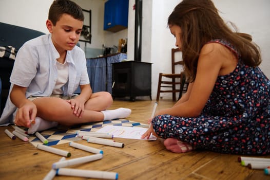 Two children are seated on the floor engaged in drawing and coloring activities. Art supplies are spread around, creating a cozy and creative atmosphere.