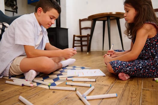 Two children engaging in creative drawing while sitting on the floor at home, surrounded by colorful markers. A warm and relaxed environment encouraging imagination and teamwork.