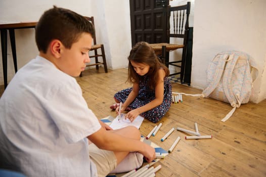 Two children engaging in drawing activities on a rustic wooden floor, surrounded by art supplies and a backpack, creating a collaborative and creative atmosphere.