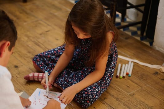 Two children engaged in artistic activity, drawing with markers on paper, conveying creativity and childhood bonding. The wooden floor adds a warm and homely environment to the scene.