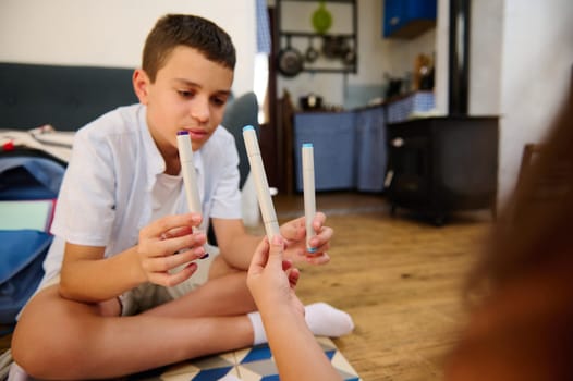 A boy focuses while holding markers, sitting on a wooden floor, engaging in an artistic activity in a cozy home setting.