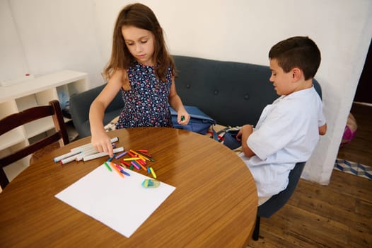 Two children sit at a wooden table working on creative crafts with markers, pencils, and paper. The scene emphasizes creativity, childhood learning, and the joy of art-making activities.