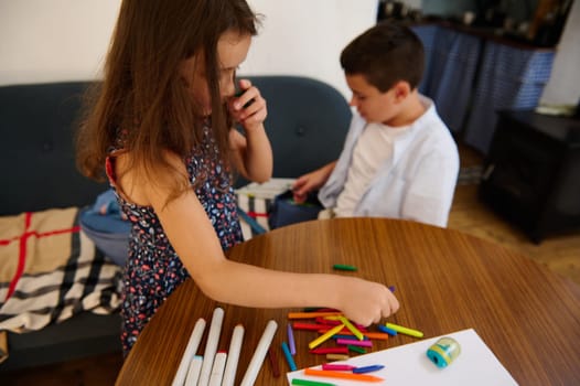 A young girl and boy are engaged in creative activities, arranging and selecting drawing tools on a wooden table in a comfortable, homey setting.