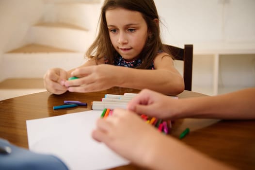 Young girl sitting at a table drawing with colored markers, expressing creativity and focused on her activity.