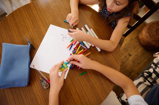 Two children are sitting at a wooden table, creating art with colorful markers and paper, capturing creativity and collaboration.