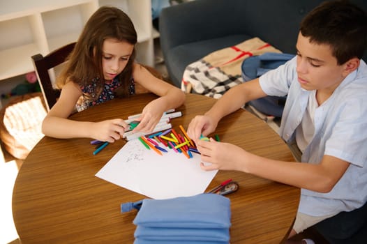 Two children sit at a table engrossed in a collaborative creative activity, sorting colorful markers and making art on blank paper.