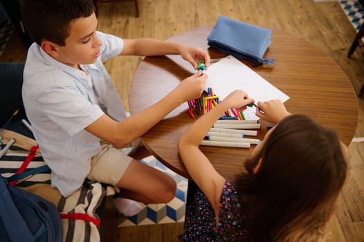 Two children seated at a wooden table organizing markers. The kids are immersed in their creative activity, with a focus on collaboration and artistic preparation. A casual and educational moment.