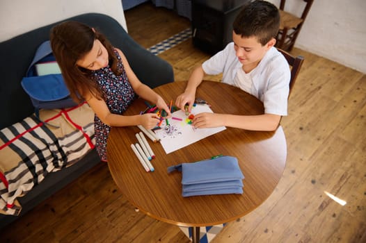 Two children drawing and coloring on paper at a small wooden table in a cozy room.
