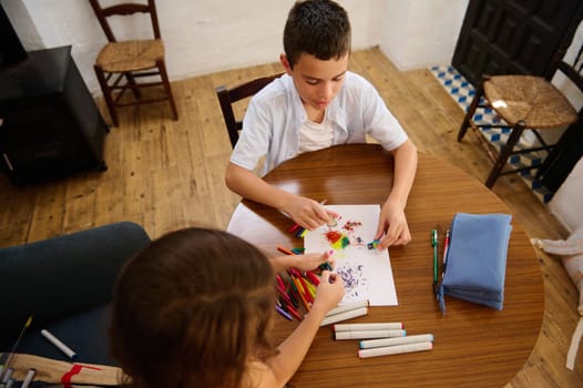 Two children sitting at a table with art supplies, drawing colorful artwork with vibrant markers in a creative and cozy indoor environment.