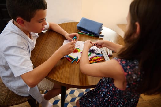 Two children sit at a table engaged in creative artistic activities using crayons and paper.