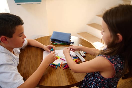 Two children sitting at a round table, immersed in artistic activity using crayons and paper, fostering creativity and collaboration in a cozy home setting.