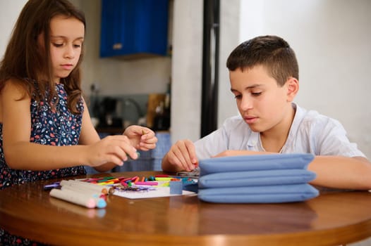Two children creating artwork with colored materials on a table, showcasing creativity and focus in a home setting.