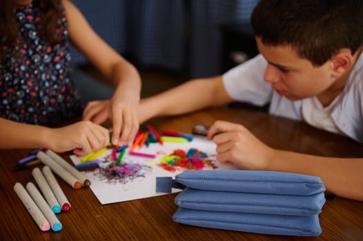 Two kids collaborating on an art project using crayons and markers. Hands engaging in creativity and art materials on a table. Depicts teamwork, imagination, and the joy of artistic expression.