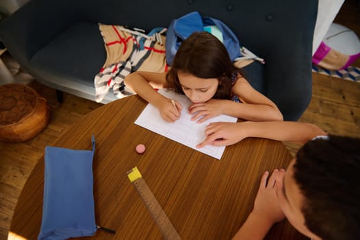 Young girl focuses on writing with help from an adult, captured over a table at home, emphasizing learning.
