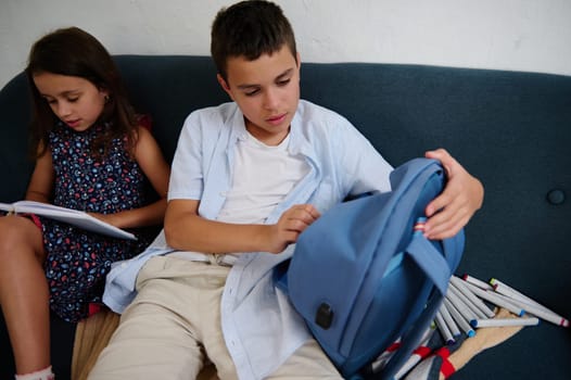 A boy and girl sitting on a couch, one organizing a backpack while the other reads and writes.