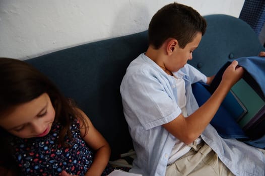 Two children sitting on a sofa, one looking into a backpack. Their expressions suggest curiosity and engagement, capturing a candid moment of childhood discovery and interaction.