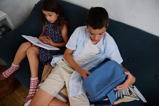 Two children, a girl reading and a boy organizing his backpack, sitting on a couch. The scene portrays focus and interaction in a casual, indoor setting.