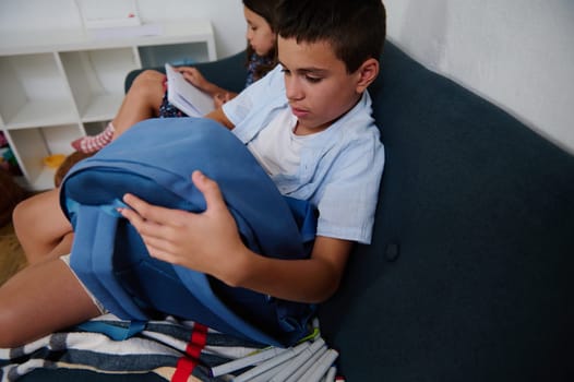 Two children engage in educational activities at home, one examining a backpack while the other reads.
