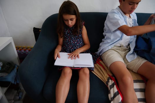 A young girl reads a book on a couch while a boy organizes his belongings. The setting conveys focus and productivity, depicting childhood learning and preparation in a household environment.