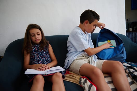 Two children sitting on a couch, one reading a book while the other organizes items in a blue backpack. They seem focused and busy with learning activities.