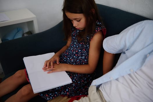 A young girl sits on a couch, intently reading from a book while being accompanied by an adult.