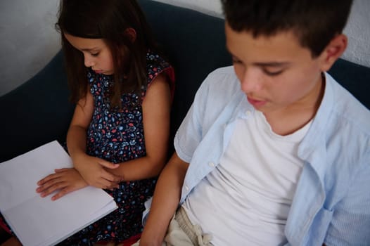 Two children are seated on a sofa, with the young girl reading a book, in a quiet indoor setting.