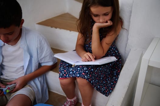 Two children engaged in reading and creative activities, enjoying learning on stairs in a cozy interior setting. A girl reads a book while a boy holds colorful markers.