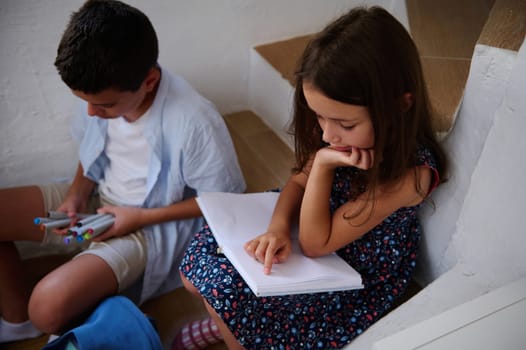 Two children are shown studying. The girl is reading from a notebook, and the boy is holding markers while seated. The environment suggests learning and focus.