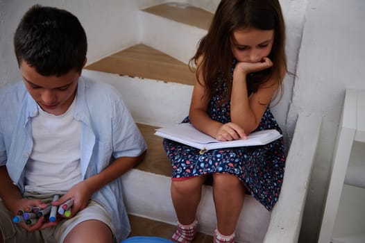 Two children sitting on stairs, one drawing with markers and the other reading a notebook, focusing on their activities.