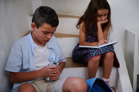 Two children sitting on stairs. The boy is placing art supplies into a bag, while the girl reads attentively from a notebook.