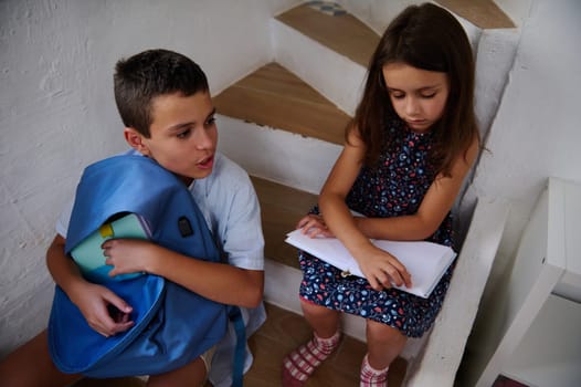 Two children sit on stairs, studying and sharing materials. They display focus and collaboration while working with books, emphasizing education and teamwork.