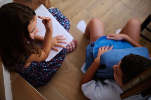 Overhead view of two children sitting on the floor, discussing notes, one holding a notebook, the other a backpack, creating a scene of youthful interaction and collaboration.