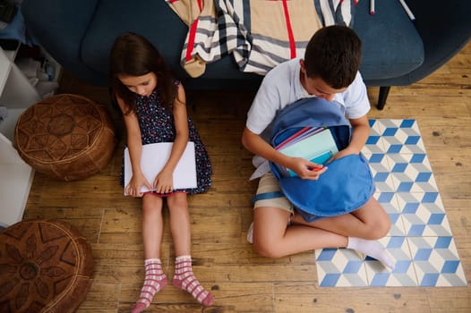 Two children sit on the floor, one organizing books while the other reads a notebook. The scene is set in a cozy indoor space with a comfortable and relaxed atmosphere.