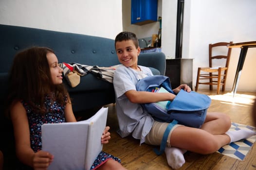 A brother and sister studying and interacting while sitting in a cozy home environment with books and backpacks.