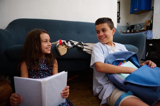 Two children sit on the floor near a sofa, smiling and interacting together, one with a book and the other with school supplies, creating a warm and friendly study moment.
