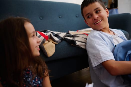 Two kids share a lighthearted moment, sitting indoors near a cozy couch with a blanket.