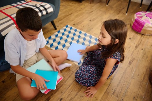 Two children sit on the floor at home, engaged in studying together with notebooks. The setting conveys education, collaboration, and a comfortable domestic environment.