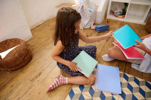 Two children sitting on a wooden floor, exchanging and arranging bright-colored notebooks within an organized and cozy environment.