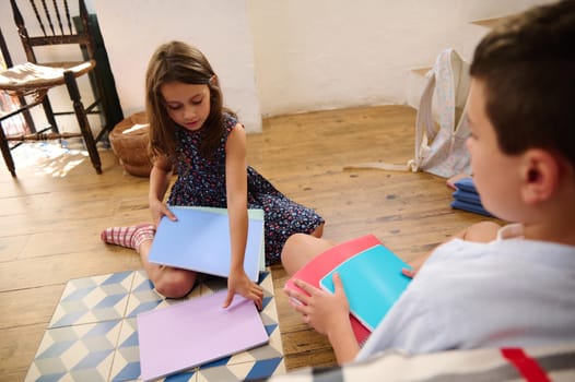 Two children sitting on a wooden floor and looking at colorful notebooks, showcasing a bright and creative learning environment.