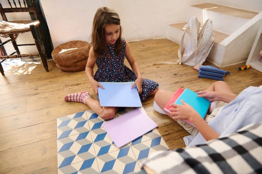 Two children sit on a wooden floor reading and studying colorful notebooks in a cozy and bright home environment.