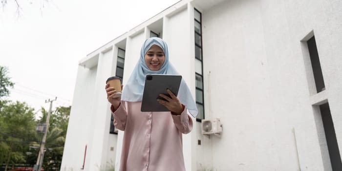 A Muslim businesswoman in a hijab smiles as she uses a tablet and holds a coffee cup, enjoying her work in an outdoor professional setting.