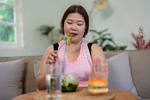 An overweight woman thoughtfully eating salad while rejecting a burger, showcasing healthy eating decisions for weight loss in a cozy home environment.