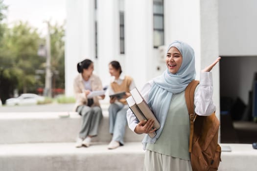 A diverse female student smiling and waving while holding books, with classmates engaged in a study session in the background on campus.
