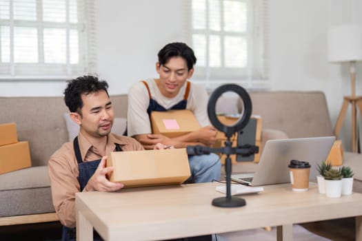 Two young Asian entrepreneurs getting ready for an unboxing session in their home office, holding packages and setting up equipment to engage with their audience.