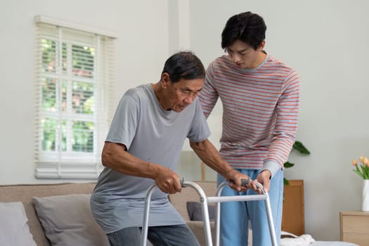 A caring son supports his elderly father as he uses a walker in their bright living room, illustrating the bond and dedication of family care.