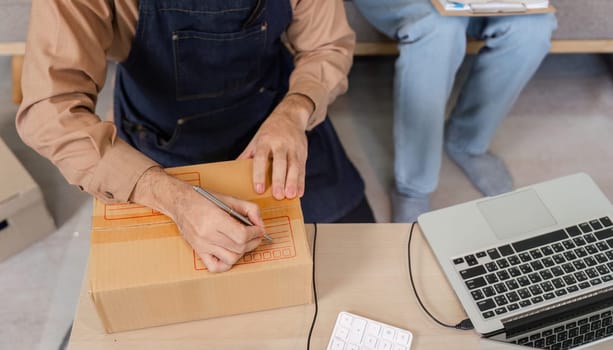 A focused social media marketer labeling product packages with a pen while preparing for a live stream, alongside a laptop and additional packages in their home office.