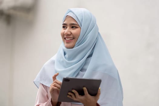 A Muslim businesswoman smiling as she holds a tablet, representing engagement and professionalism in a casual setting.