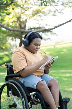 A diverse young man using a wheelchair, focused on sketching in a notebook while enjoying music with headphones in a serene outdoor environment.
