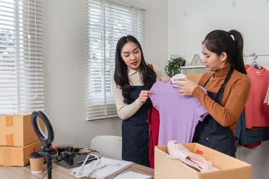 Two women engaging in a friendly discussion while presenting a vibrant purple top and red clothing, preparing for their online shop in a bright home office.