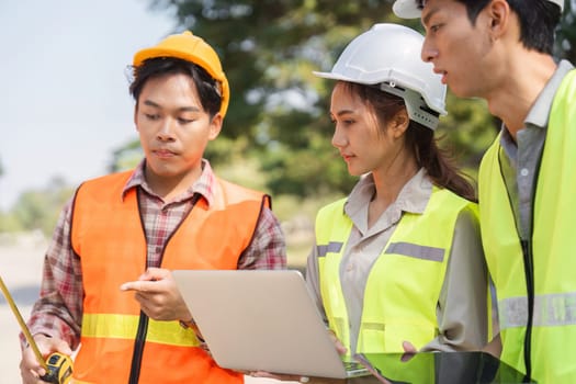 Three construction workers, including an engineer and a supervisor, are analyzing plans and discussing measurements on-site while ensuring safety with helmets and vests.